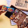 Hand holding a mini chocolate bar reading “Bun in. Oven on.” above an open selection box – a fun and thoughtful gift for mums-to-be.