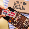 Hand holding a chocolate bar that says “Here’s to not dying alone!” in front of an open gift box – a playful chocolate gift for newly engaged couples.