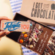 Hand holding a chocolate bar reading “I Tolerate You” above an open gift box – a playful choice for couples with a sense of humour.