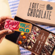 Hand holding a chocolate bar that reads “Sorry You Got Old” in front of an open birthday gift box – filled with five handmade bars featuring fun messages and toppings.