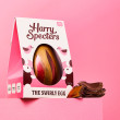 A milk chocolate Easter egg decorated with red and gold cocoa butter, next to a stack of milk chocolate Easter egg pieces