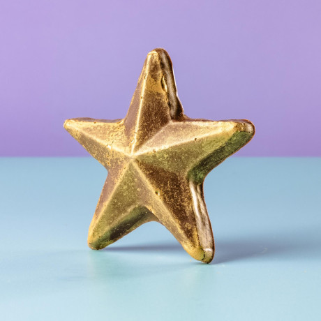 Gold shimmering chocolate star standing upright on a pastel surface – a simple reward for effort, achievement, or turning up.