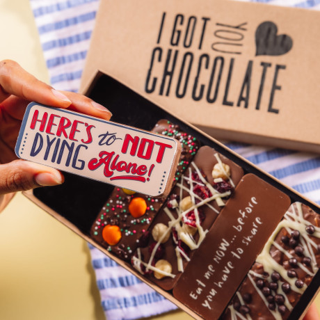 Hand holding a chocolate bar that says “Here’s to not dying alone!” in front of an open gift box – a playful chocolate gift for newly engaged couples.