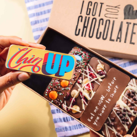 Hand holding a “Chin Up” chocolate bar in front of an open gift box – five handmade Belgian chocolate bars with playful messages and toppings, ideal for get well gifting.